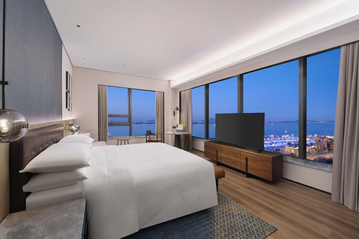 A modern hotel room with a large bed, a flat-screen TV, a desk, and a view of the city at dusk.