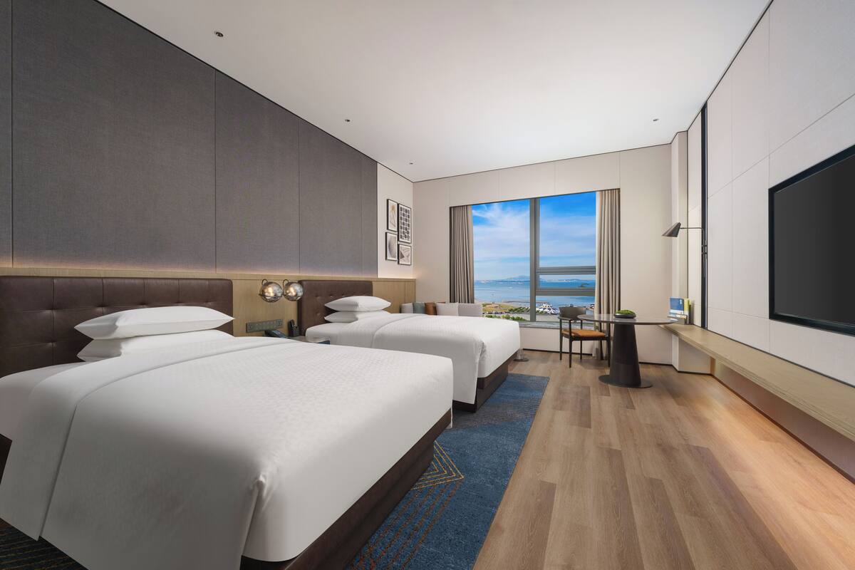 A hotel room with two beds, a desk, a TV, and a large window with a view.