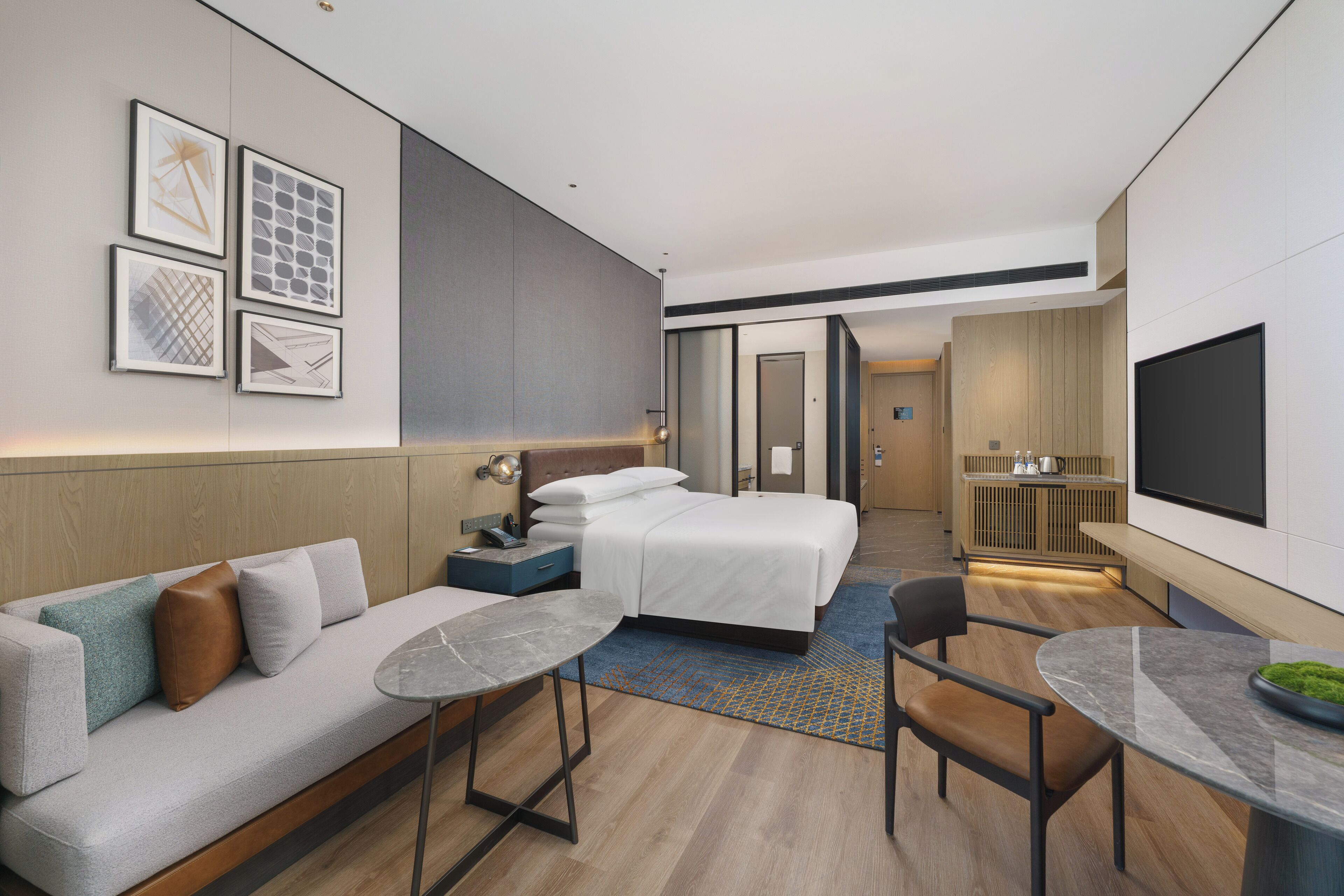 A modern hotel room with a bed, sofa, dining table, and artwork on the walls.