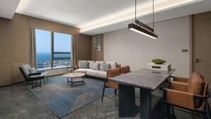 Habitación de hotel moderna con un amplio ventanal que ofrece vistas a la ciudad, un sofá, una mesa de comedor y una araña.