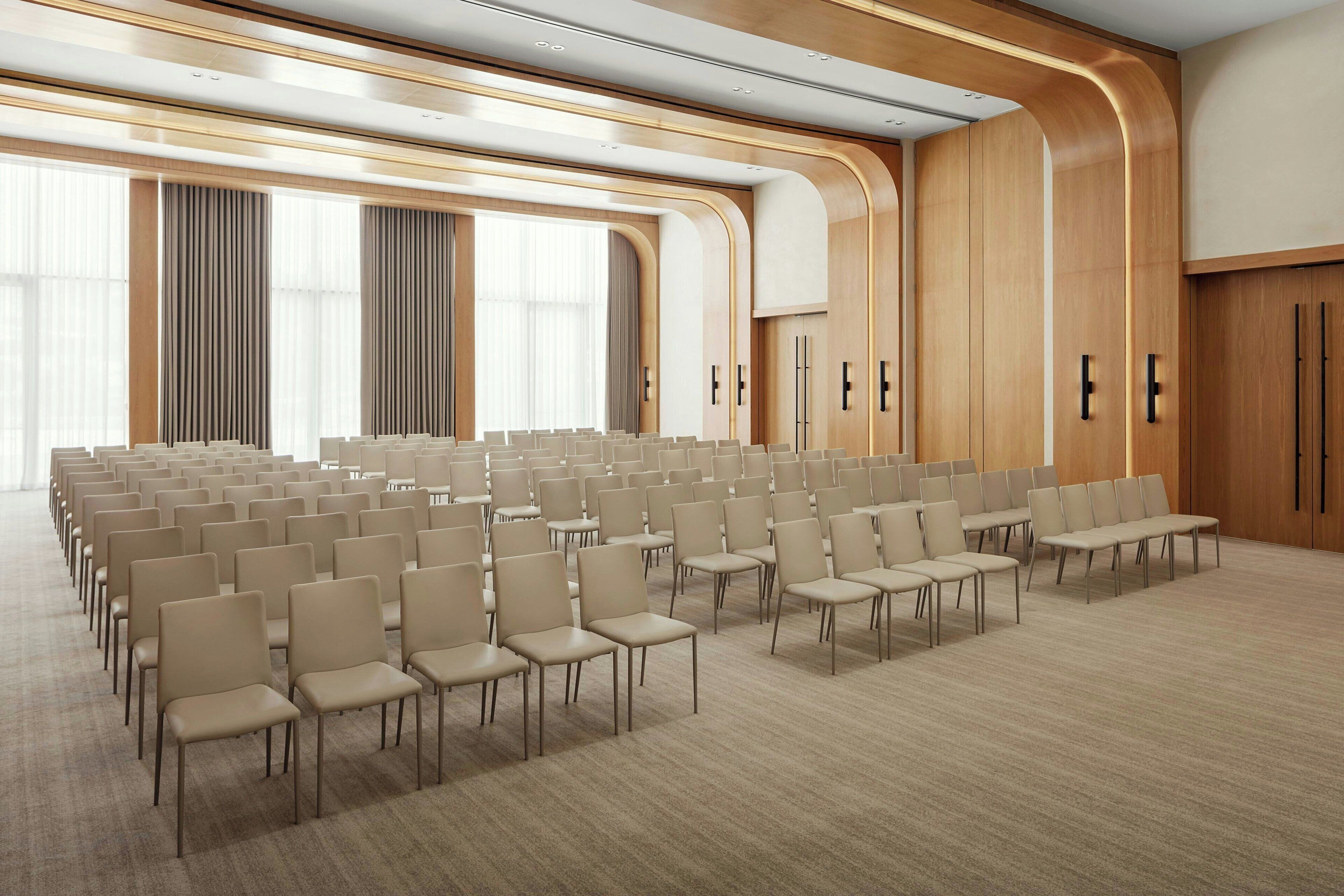 A conference room with rows of beige chairs, wooden paneling, and large windows.