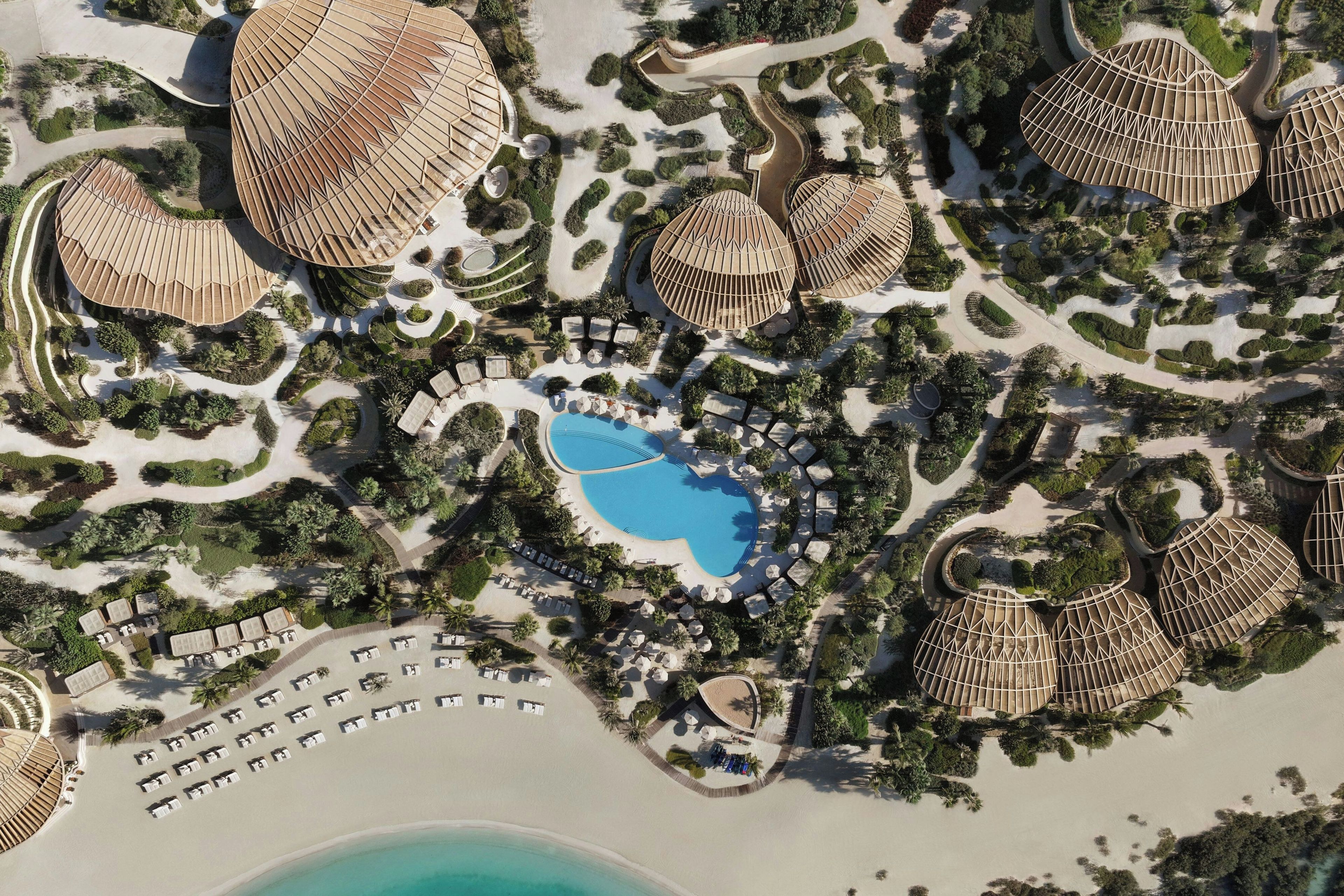 Eine Luftaufnahme eines Resorts mit mehreren kuppelförmigen Gebäuden, einem zentralen Swimmingpool und einem Strandbereich mit Liegestühlen.