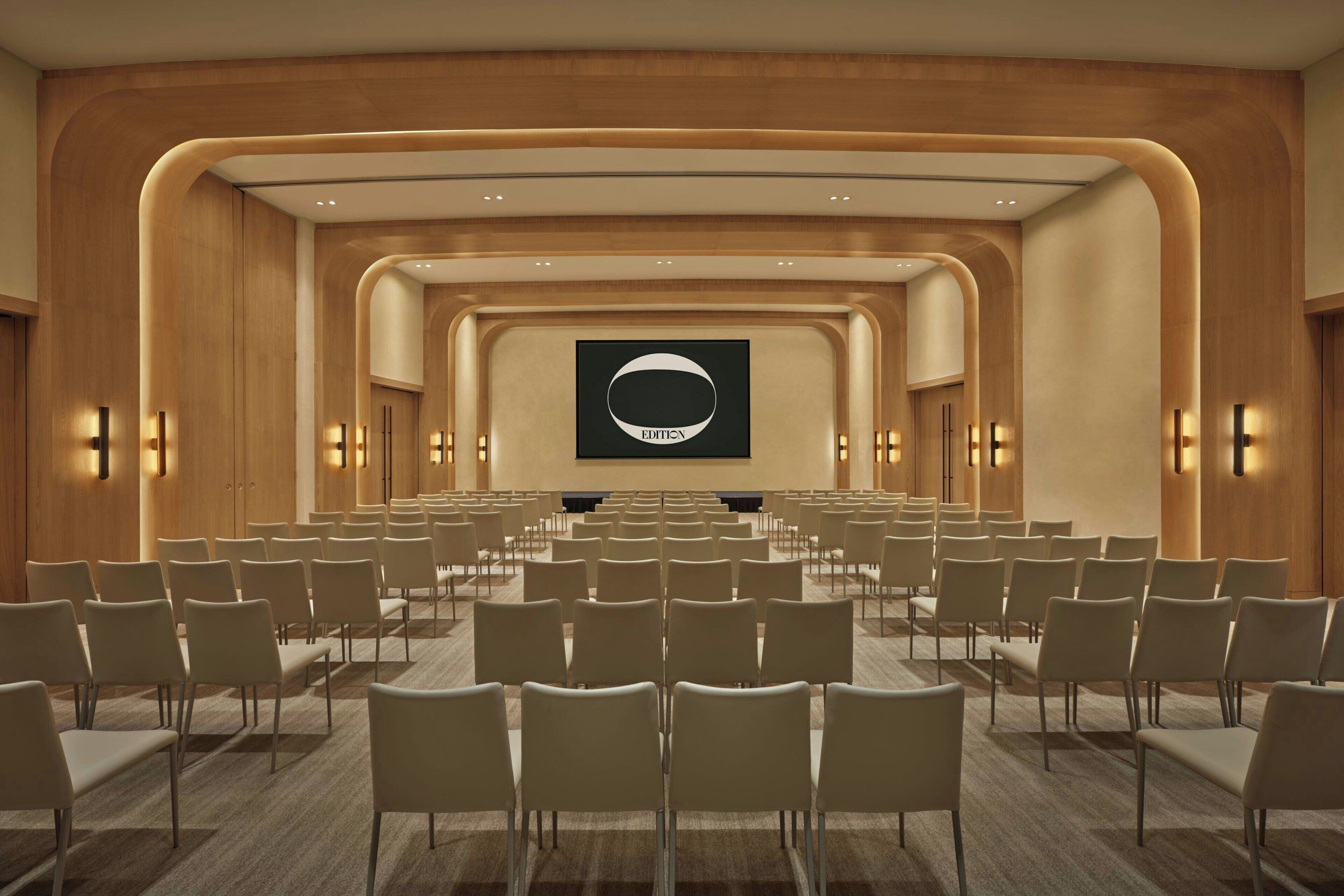 A conference room with rows of chairs, a large screen, and wall-mounted lighting.