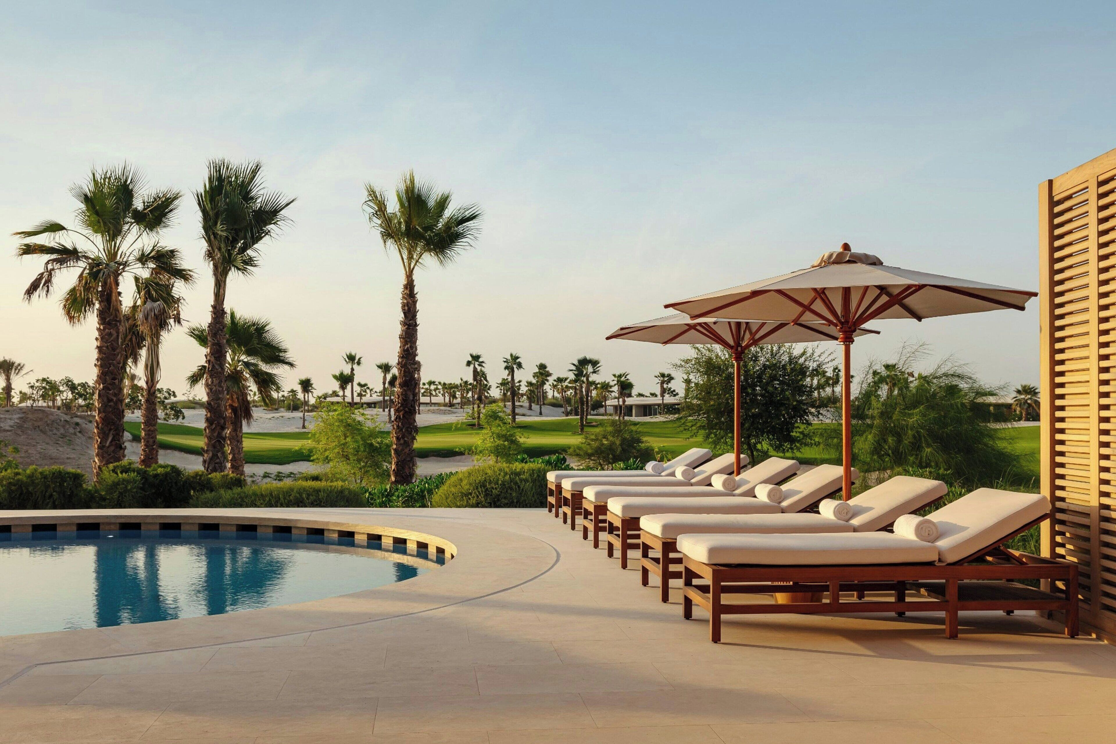 A poolside area with lounge chairs and umbrellas, palm trees, and a golf course in the background.