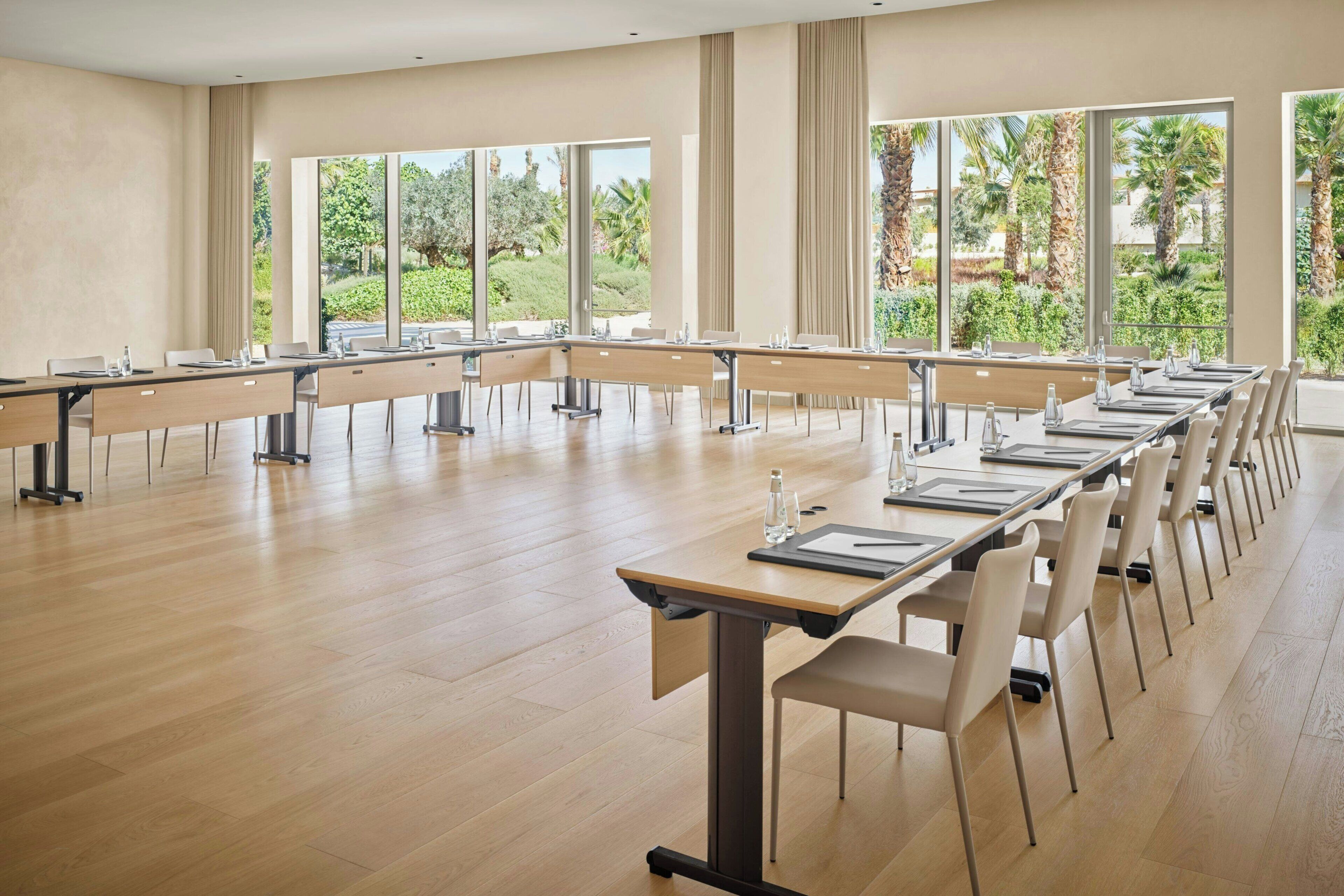 A conference room with long tables, chairs, and large windows with a view of greenery.