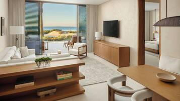 A modern living room with a sofa, dining table, and chairs, a TV mounted on the wall, and a view of the beach through large windows.