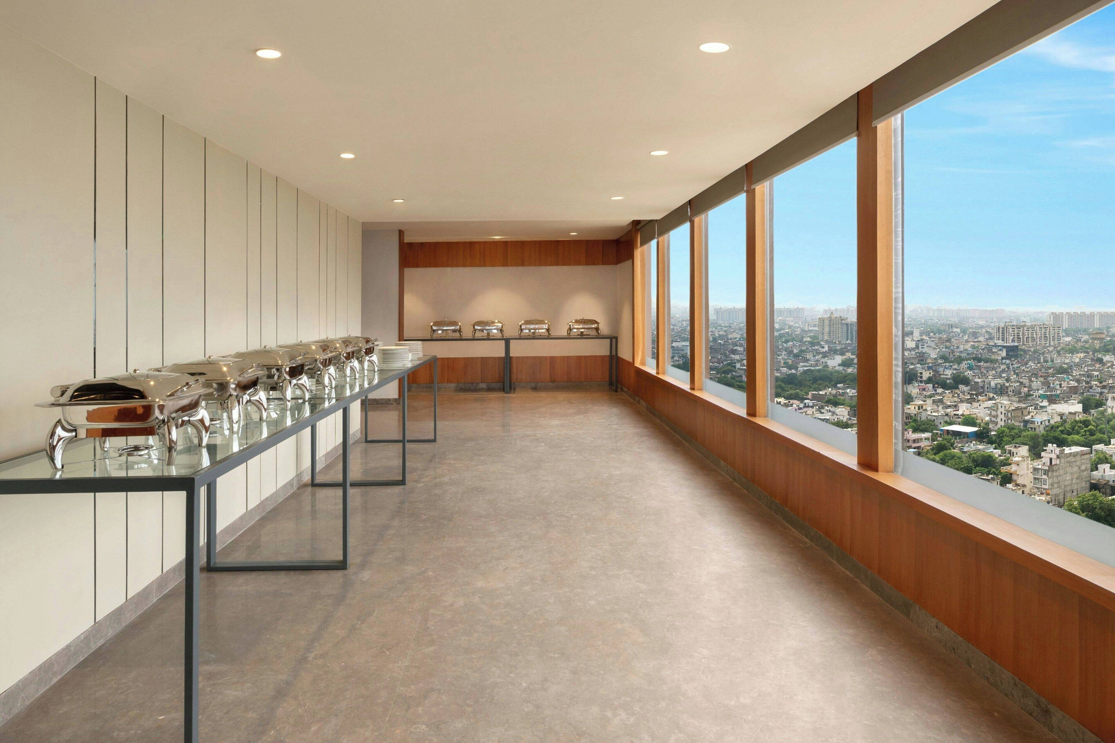 A long, narrow room with a glass table and chairs, large windows with a city view, and a high ceiling with recessed lighting.