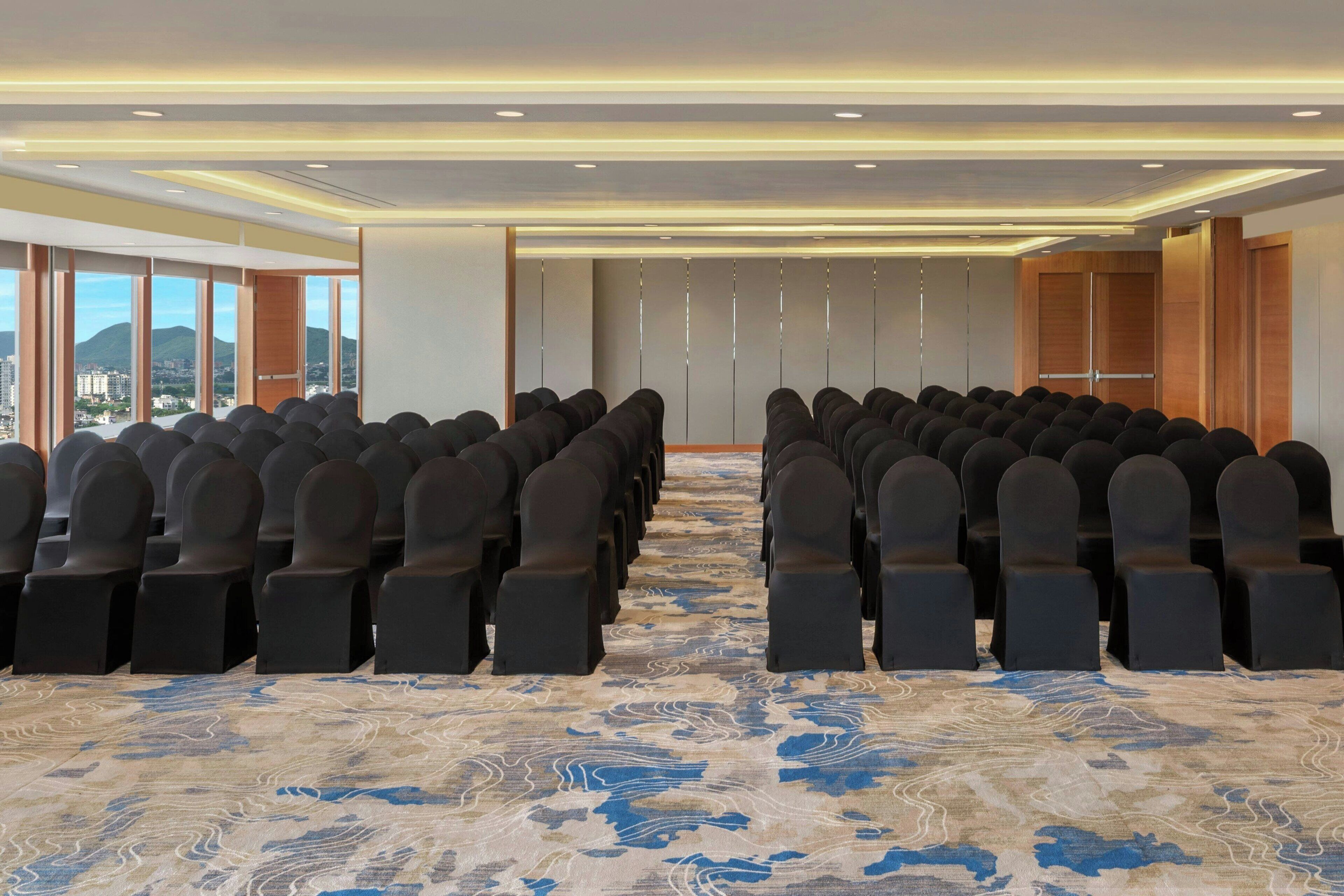 Un salón de conferencias con filas de sillas, una alfombra estampada y ventanas amplias que ofrecen vistas a edificios y montañas.