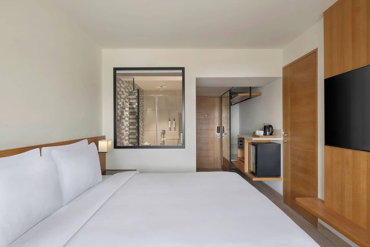 Ein Hotelzimmer mit einem großen Bett, einem Fernseher, einem Spiegel, einer kleinen Küchenzeile und einem durch eine offene Tür einsehbaren Badezimmer.
