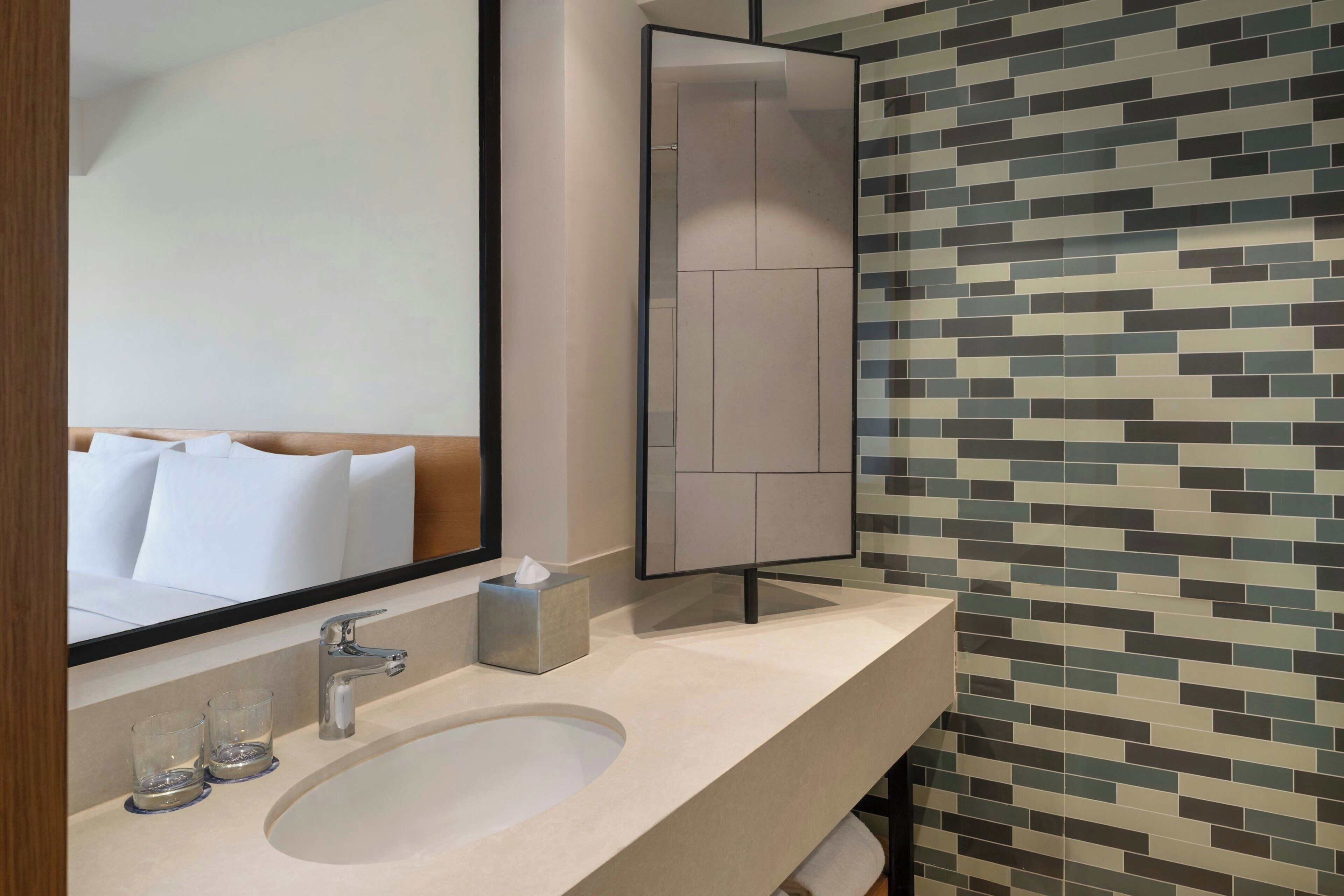 A modern bathroom with a large mirror, a sink, two glasses, a towel, and a tiled wall.