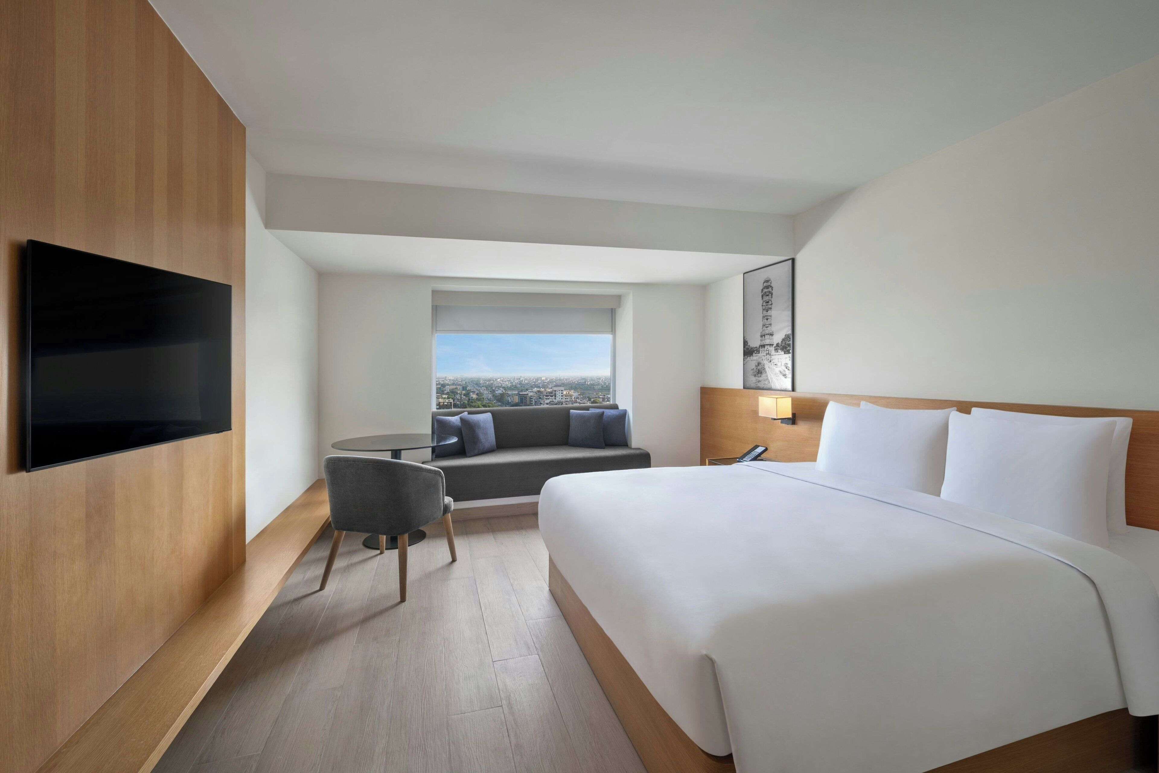 Habitación de hotel con una cama grande, un televisor de pantalla plana en la pared, un área de estar con un sofá y una silla, y una ventana con vistas a la ciudad.