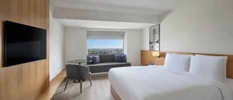 Habitación de hotel con una cama grande, un televisor de pantalla plana en la pared, un área de estar con un sofá y una silla, y una ventana con vistas a la ciudad.