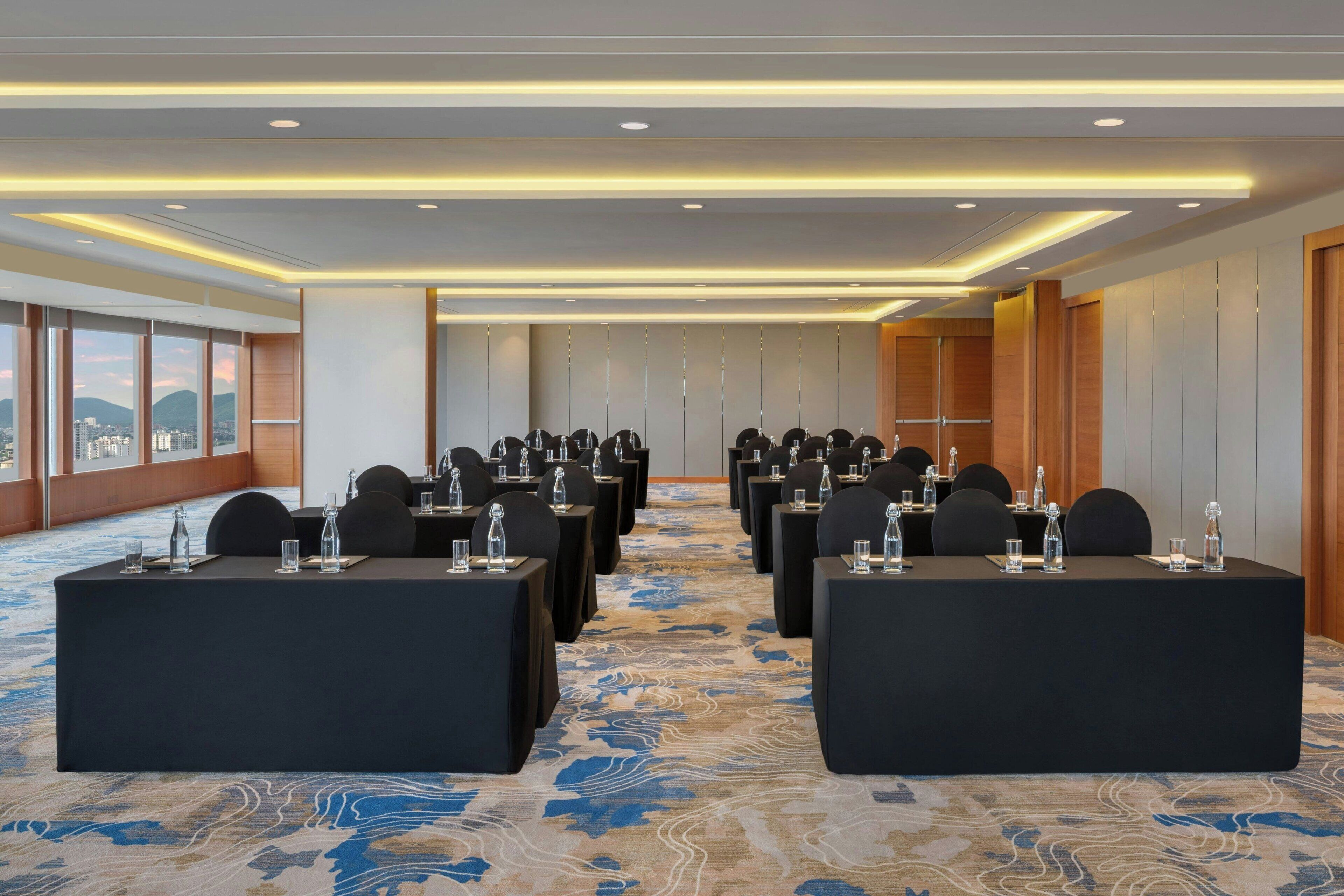 Un salón de conferencias con filas de mesas y sillas, ventanas amplias con vista a la ciudad y una alfombra estampada.