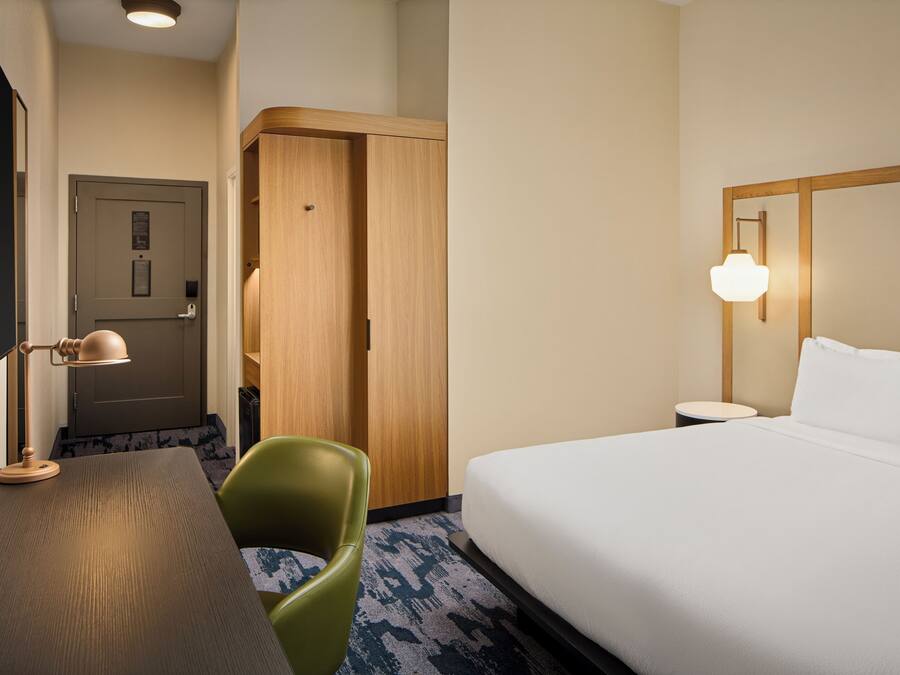 Habitación de hotel con cama, escritorio, silla, armario y mesita de noche con lámpara.