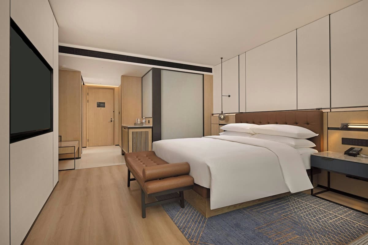 A modern hotel room with a large bed, a desk, a bench, and a walk-in closet.
