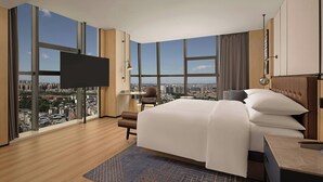 Quarto de hotel moderno com uma cama grande, televisão de tela plana, escrivaninha e vista para a cidade através de amplas janelas.