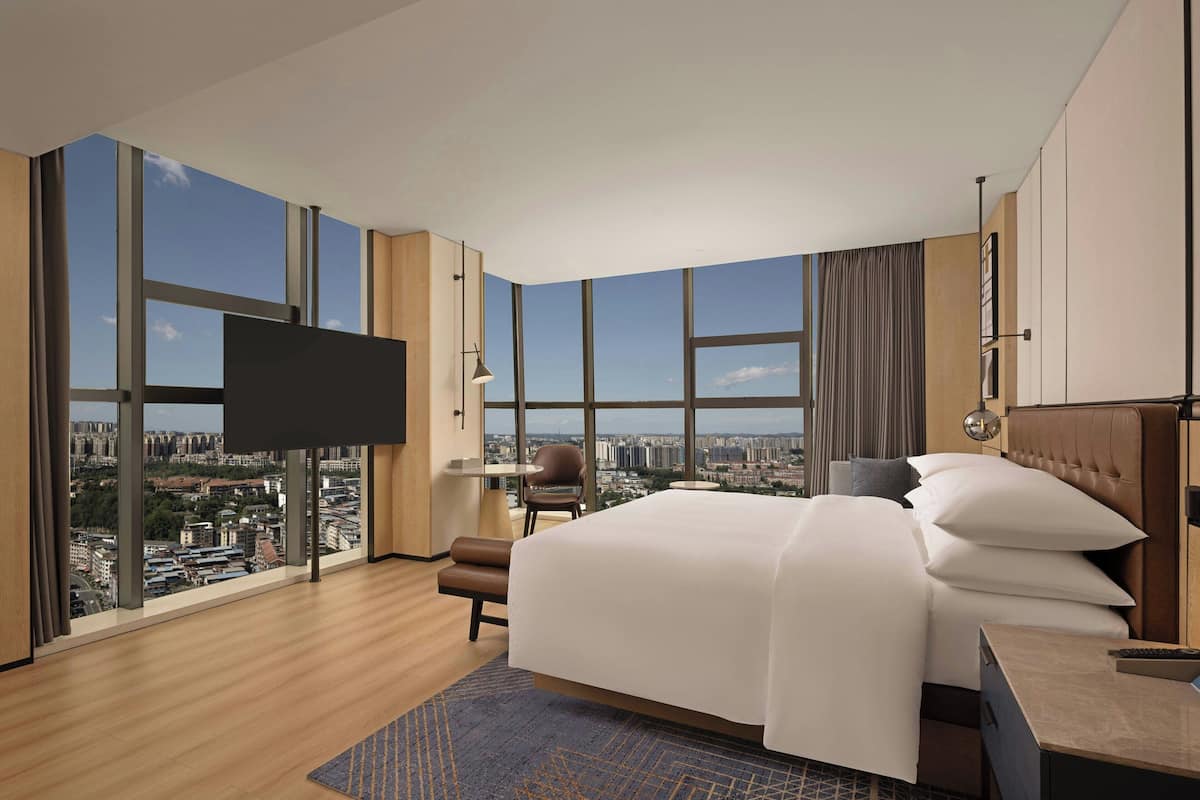 A modern hotel room with a large bed, a flat-screen TV, a desk, and a view of the cityscape through large windows.
