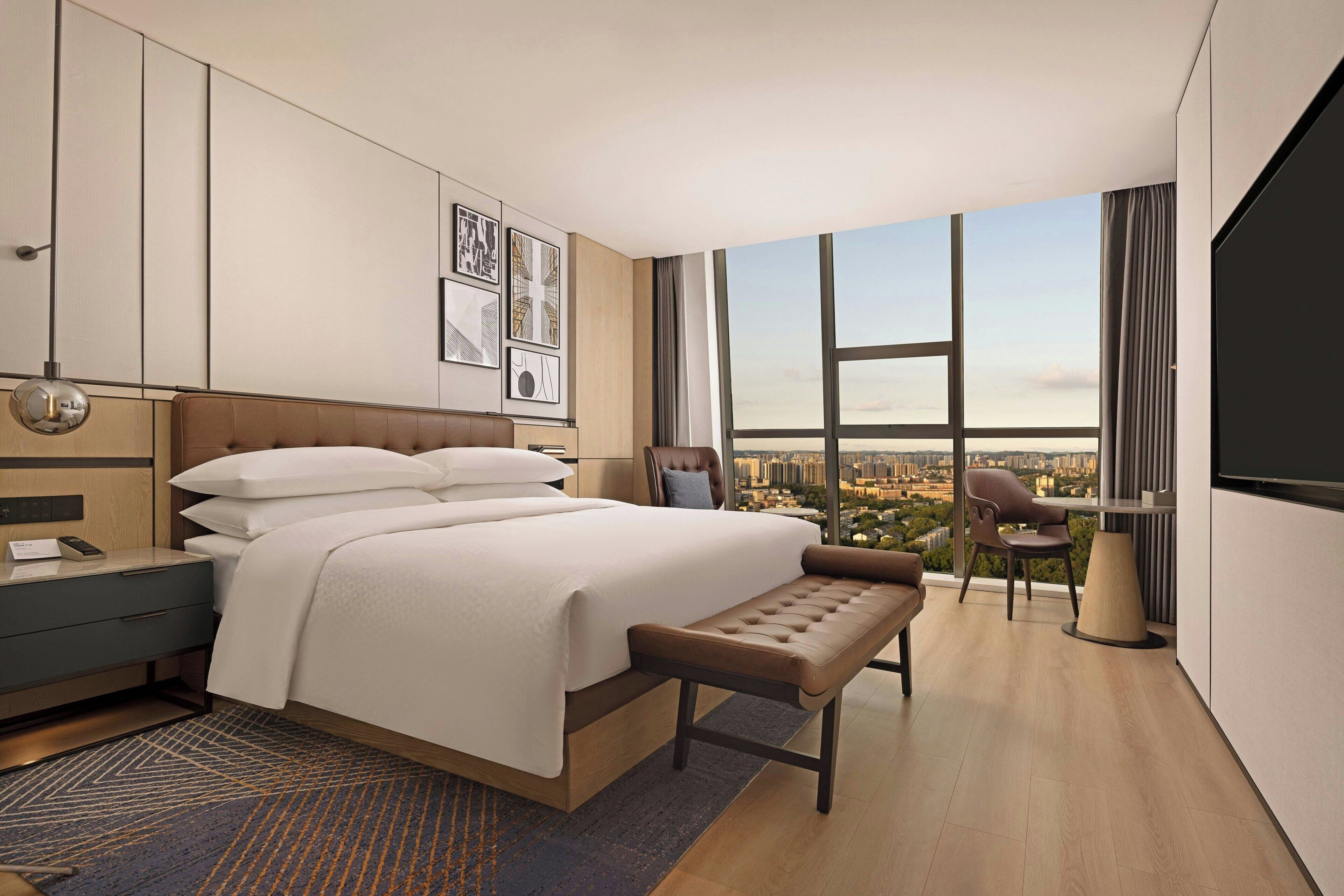 Ett modernt hotellrum med en stor säng, en bänk, ett skrivbord och en stol. Det finns ett stort fönster med utsikt över staden.