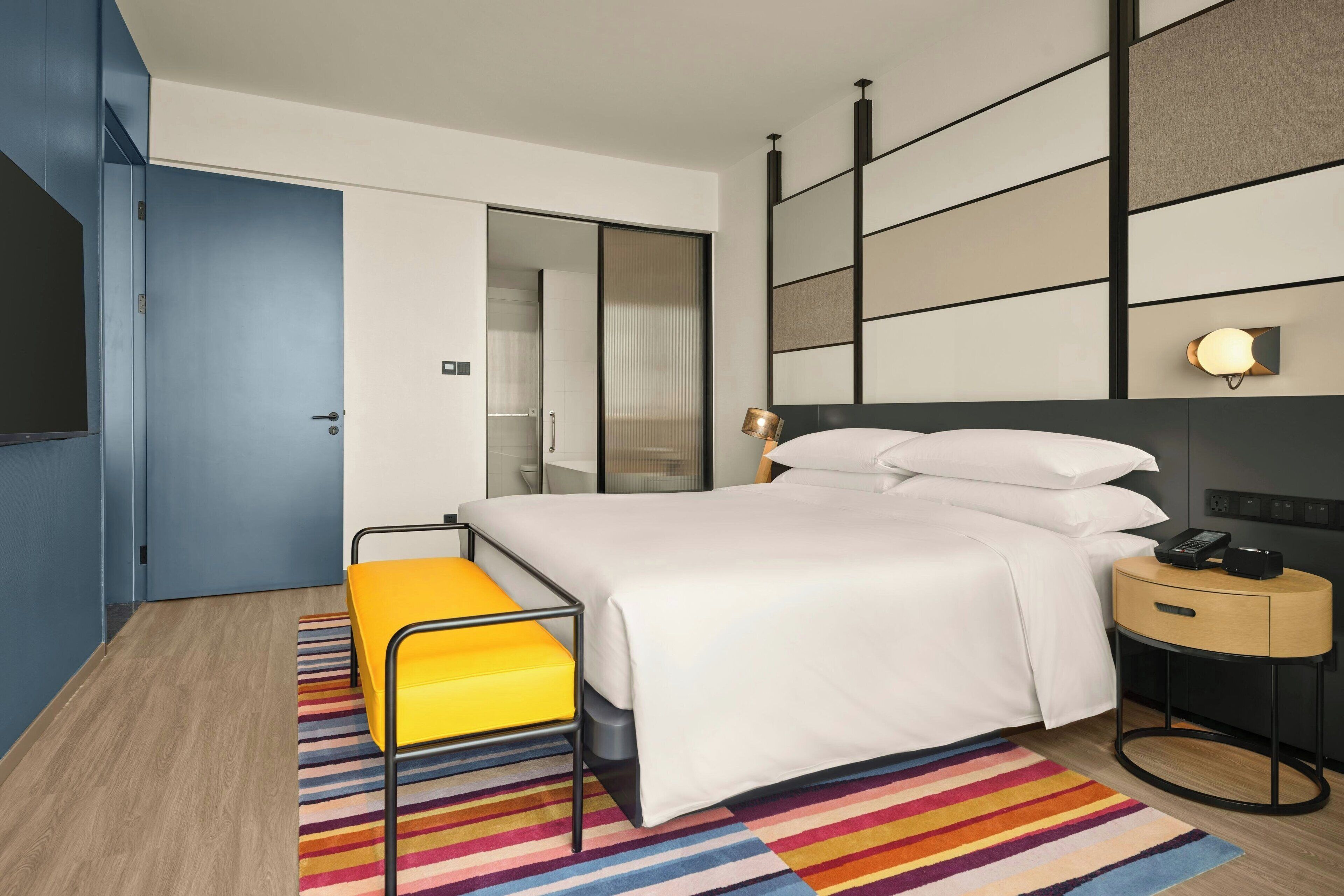 Moderni hotellihuone, jossa on sänky, yöpöytä lampun kanssa, pieni jakkara ja värikäs raidallinen matto.
