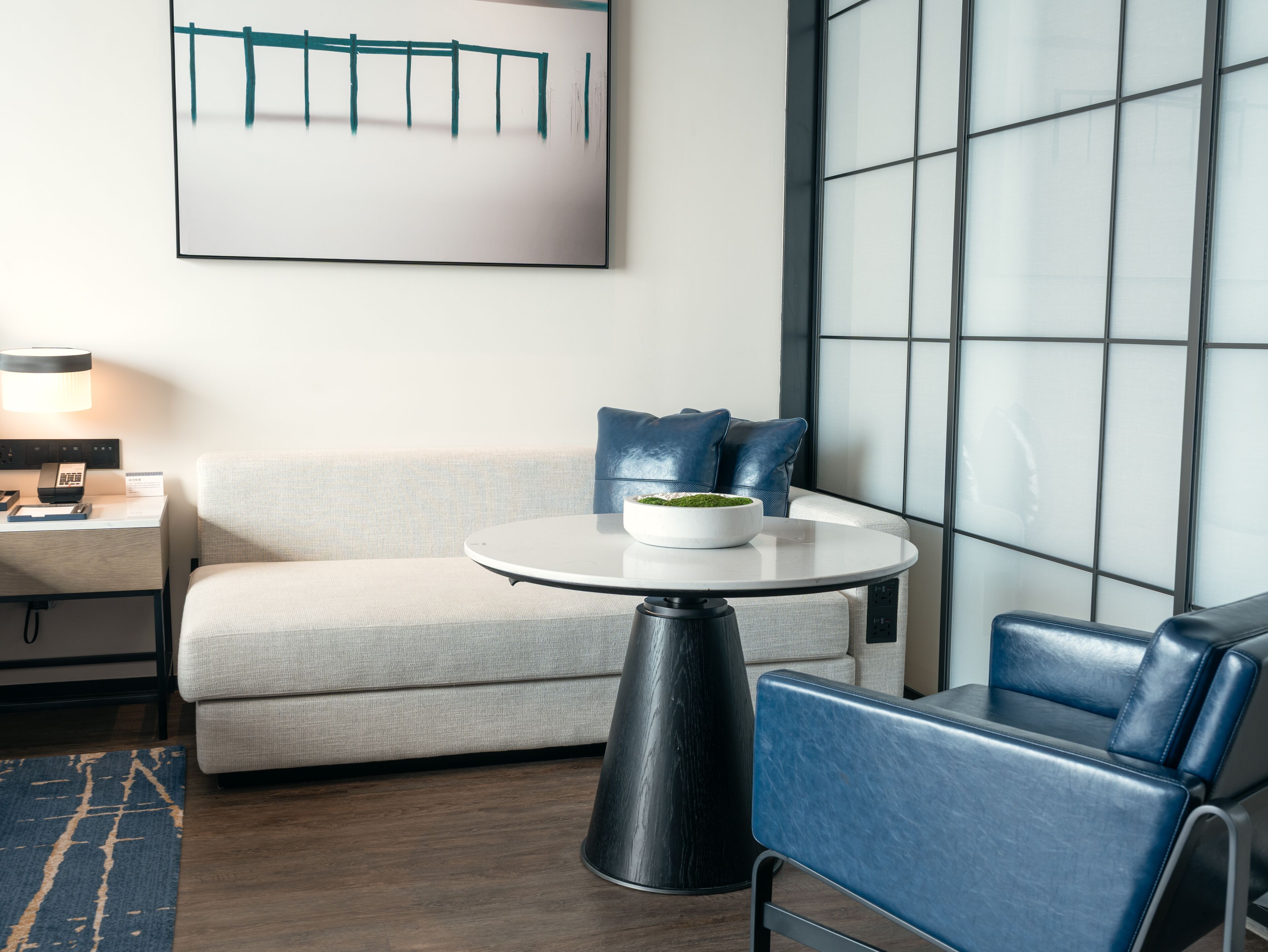 A modern hotel room with a sofa, a round table with a bowl and blue cushions, a lamp, and a large window with a grid pattern.