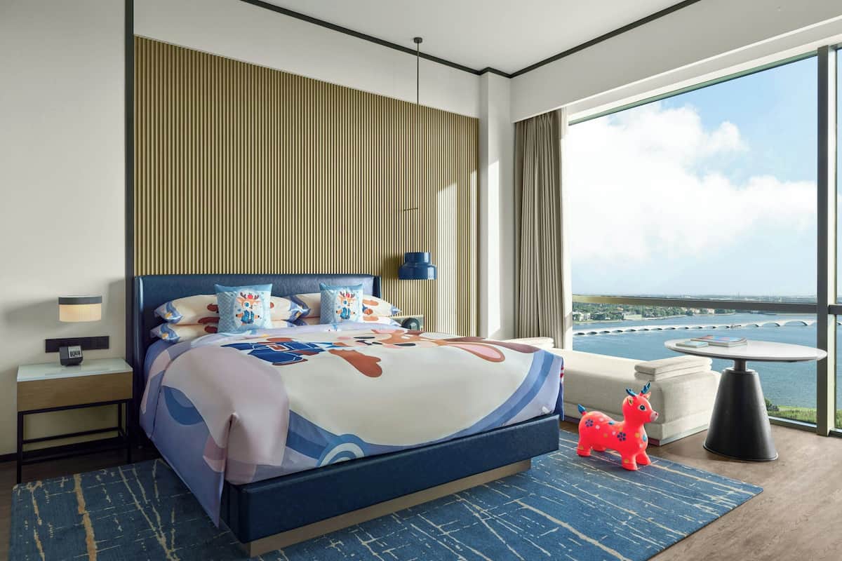 Een moderne hotelkamer met een groot bed, uitzicht op de stad, een klein rond tafeltje en een houten paardje.