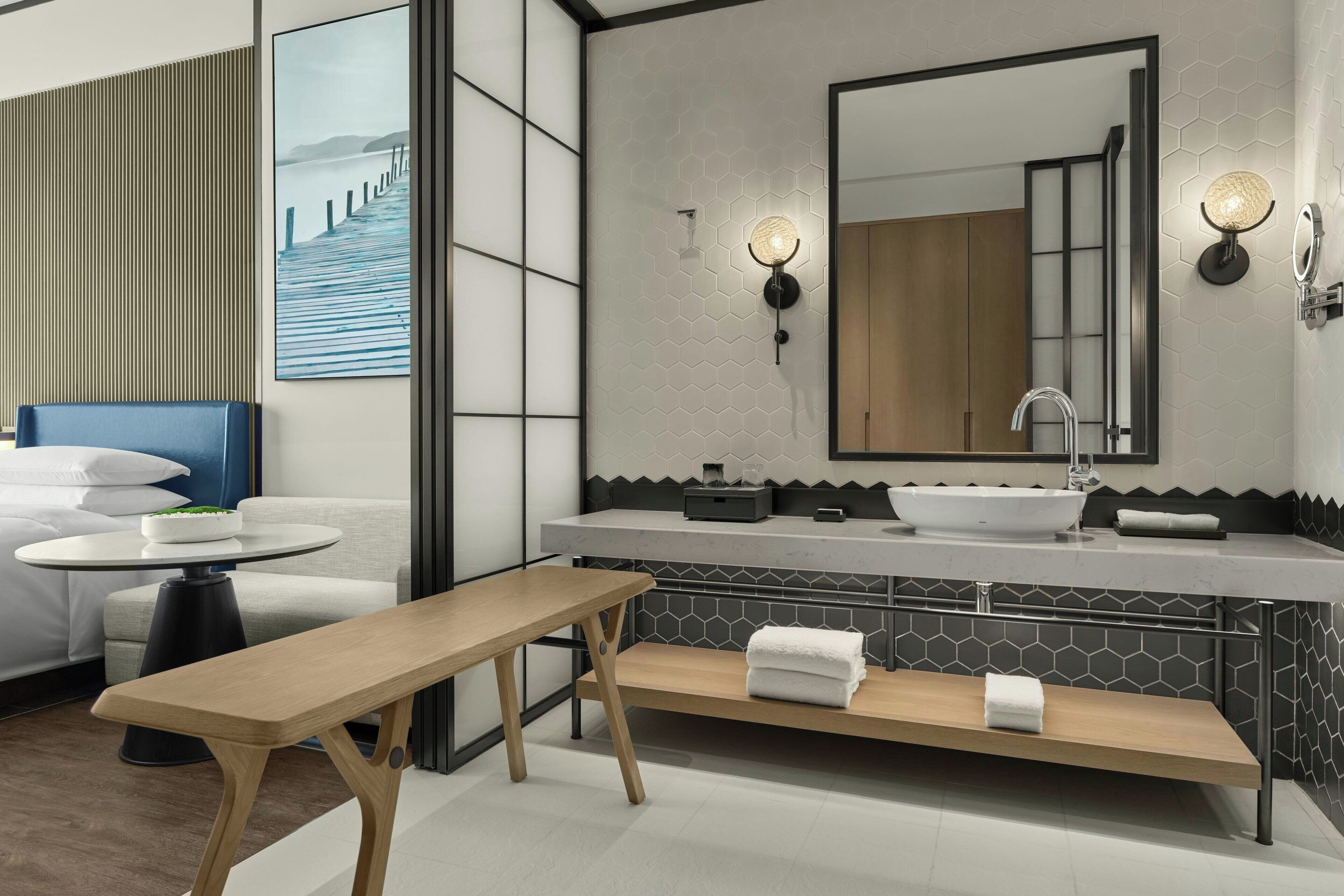 Une salle de bain moderne avec un grand miroir, un lavabo et un banc. On y trouve une table ronde sur laquelle est posé un saladier rempli de fruits, ainsi qu’une chaise bleue.