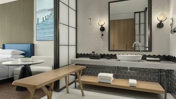 Une salle de bain moderne avec un grand miroir, un lavabo et un banc. On y trouve une table ronde sur laquelle est posé un saladier rempli de fruits, ainsi qu’une chaise bleue.