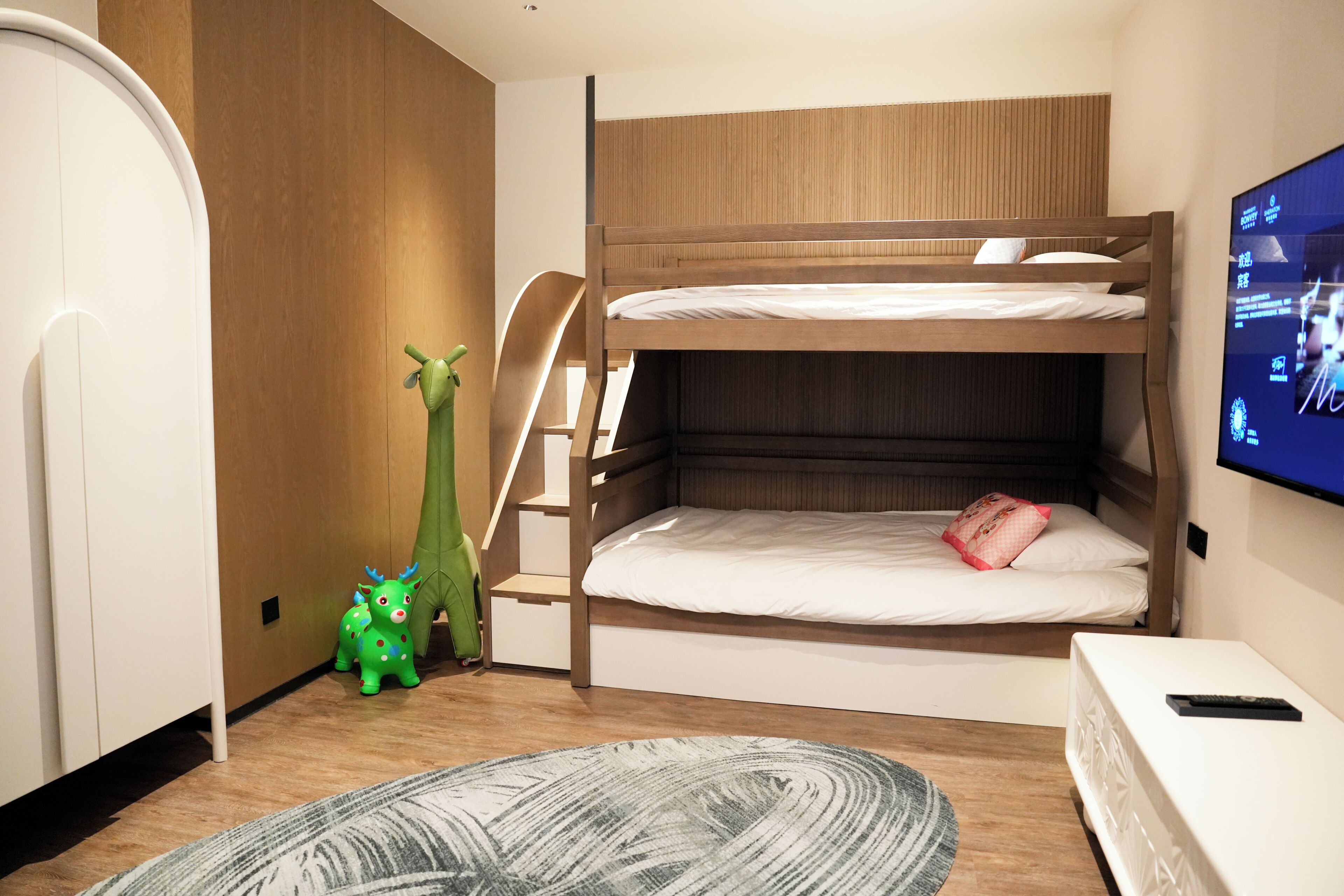 Een slaapkamer met een stapelbed, een televisie aan de muur, een groen speeltje en een kamerbreed tapijt met een patroon.