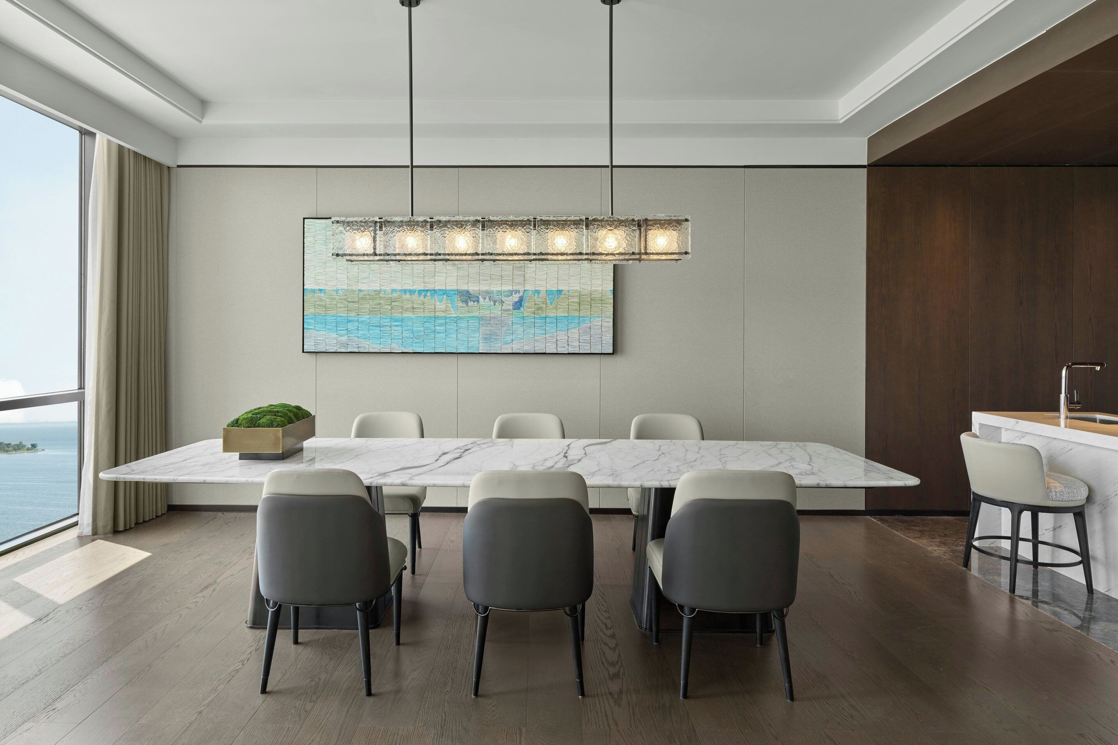 A modern dining area with a marble table, six chairs, a hanging light fixture, and a large painting on the wall.