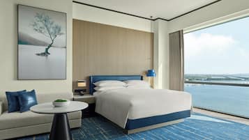 Une chambre d’hôtel moderne, dotée d’un grand lit, d’un canapé, d’une petite table, d’une table de chevet, d’une lampe et d’une grande fenêtre donnant sur un plan d’eau.