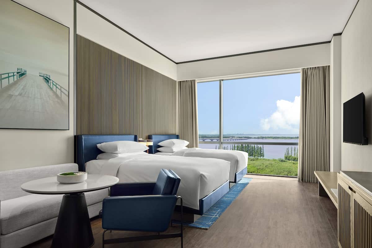 Hotelkamer met een groot bed, een bank, een kleine tafel en een televisie. De kamer heeft een groot raam met uitzicht op een waterpartij en begroeiing.