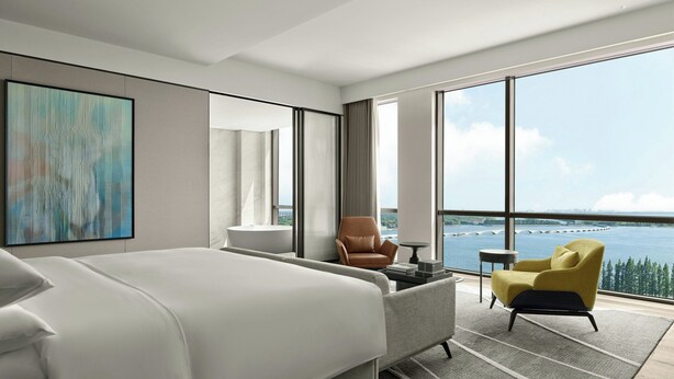 Habitación de hotel moderna con una cama grande, un área de descanso con una silla y una mesita pequeña, y un ventanal con vistas al mar.