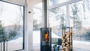 A modern room with a wood-burning stove, a log storage rack, and a view of a snowy landscape through large windows.