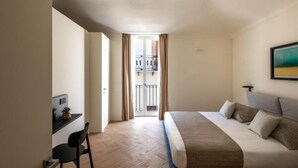 Ein Hotelzimmer mit Bett, Schreibtisch, Stuhl, einem Balkon mit Vorhängen und einem Gemälde an der Wand.