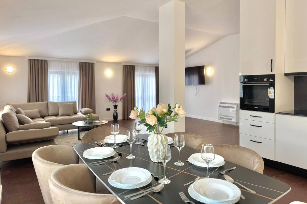 Un salon moderne avec un coin repas aménagé pour six personnes, comprenant une table noire, de l’assiette blanche et des verres à vin. On aperçoit un canapé et une table basse à l’arrière-plan.