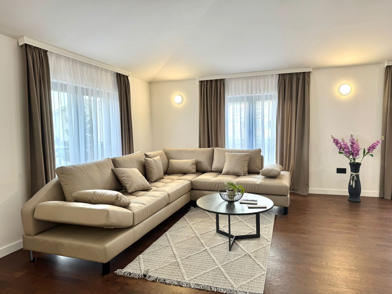 Una sala moderna con un sofá seccional color beige, una mesa de centro redonda y un jarrón decorativo con flores.