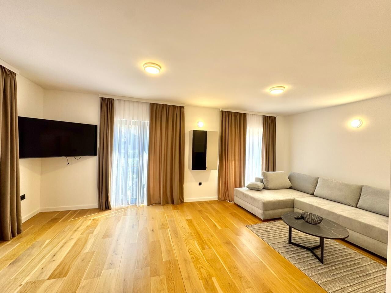 A modern living room with a large sectional sofa, a flat-screen TV mounted on the wall, and a wooden floor.