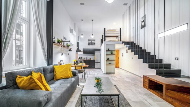 Een moderne woonkamer met een grijze bank, gele kussens, een salontafel en een trap naar een hoger gelegen verdieping.