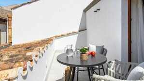 Ein Balkon mit rundem Tisch, Stühlen, einer kleinen Pflanze, einer Karaffe und einer Tomate.