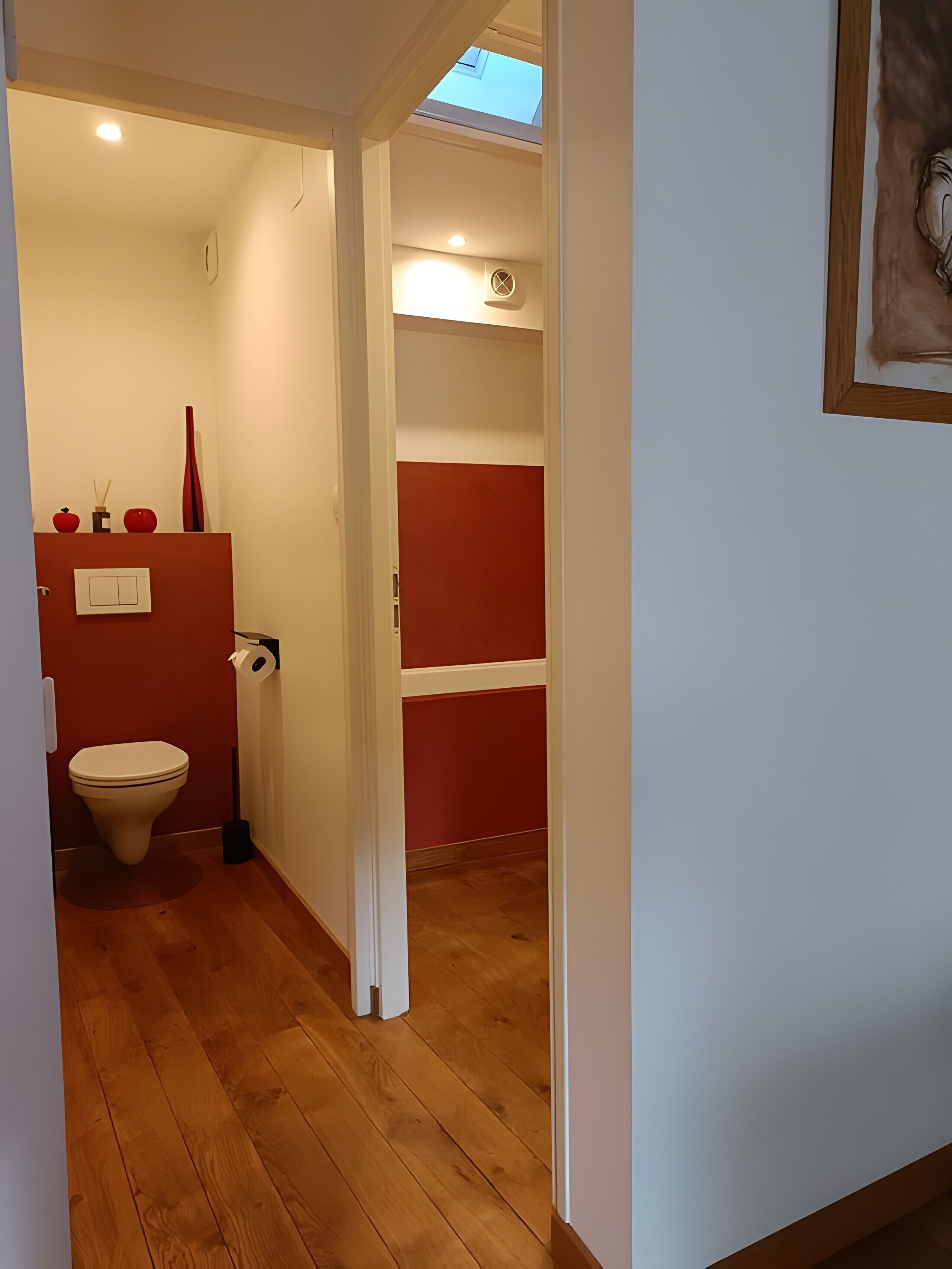 Ein schmaler Flur mit Holzboden, der zu einem Badezimmer mit roter Akzentwand und Oberlicht führt.