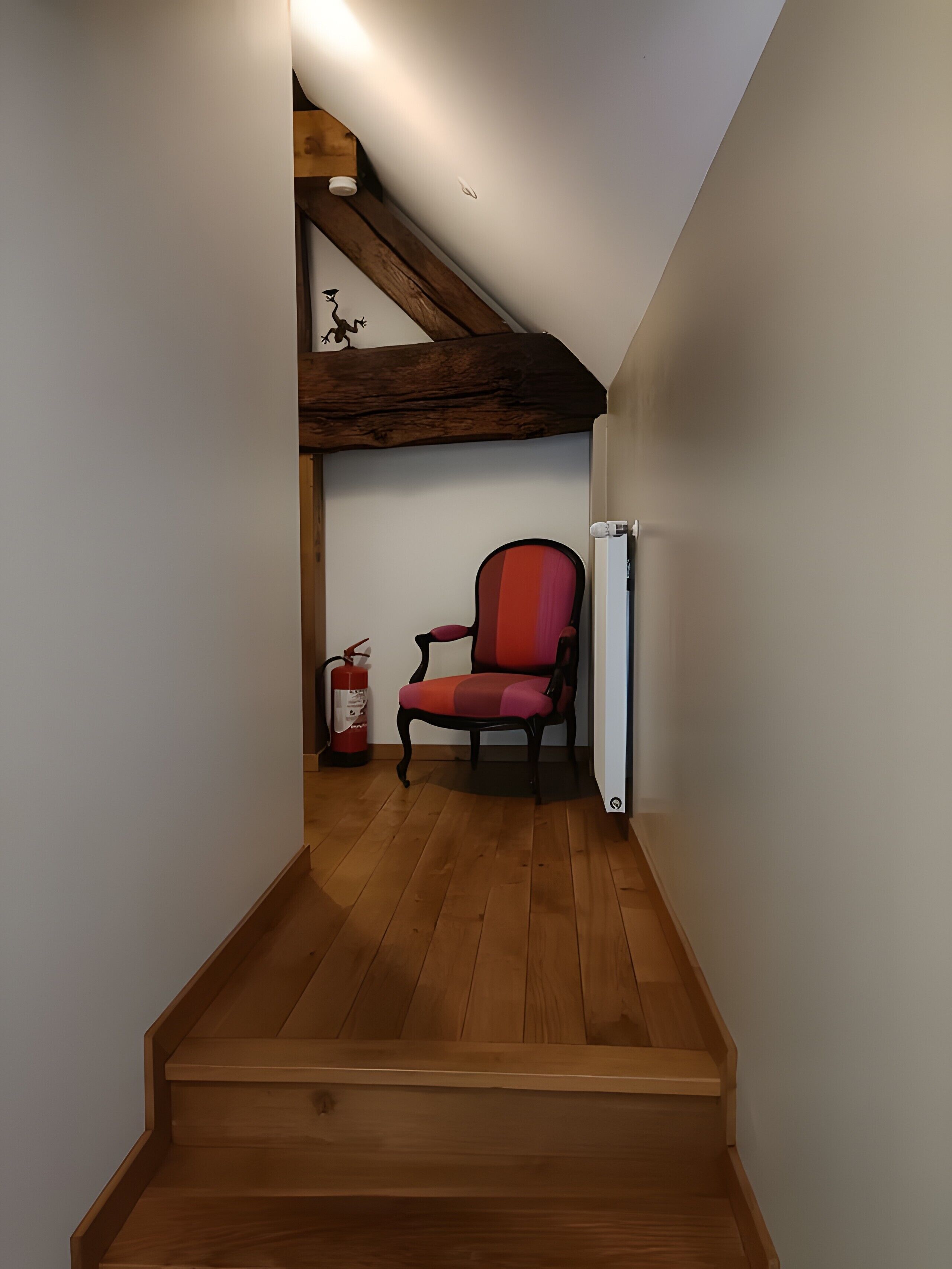 Un escalier étroit mène à une pièce où se trouvent un fauteuil rouge, des poutres en bois et un extincteur.
