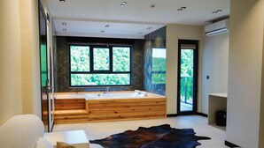 A modern bathroom with a large bathtub, wooden vanity, and a view of greenery through large windows. - Bakku House (Atakum)