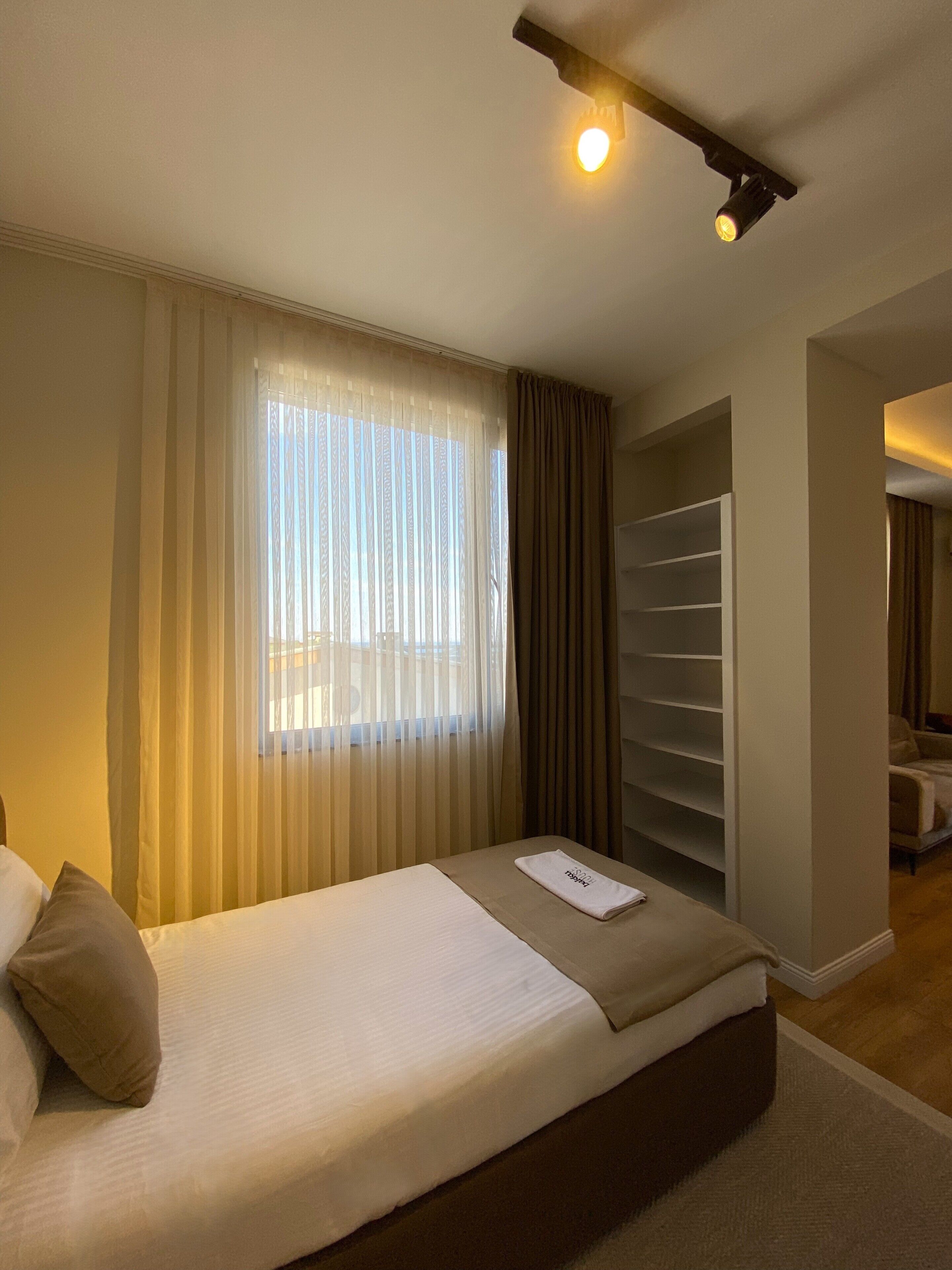 A hotel room with a bed, a window with sheer curtains, a closet with shelves, and a small table with a towel.