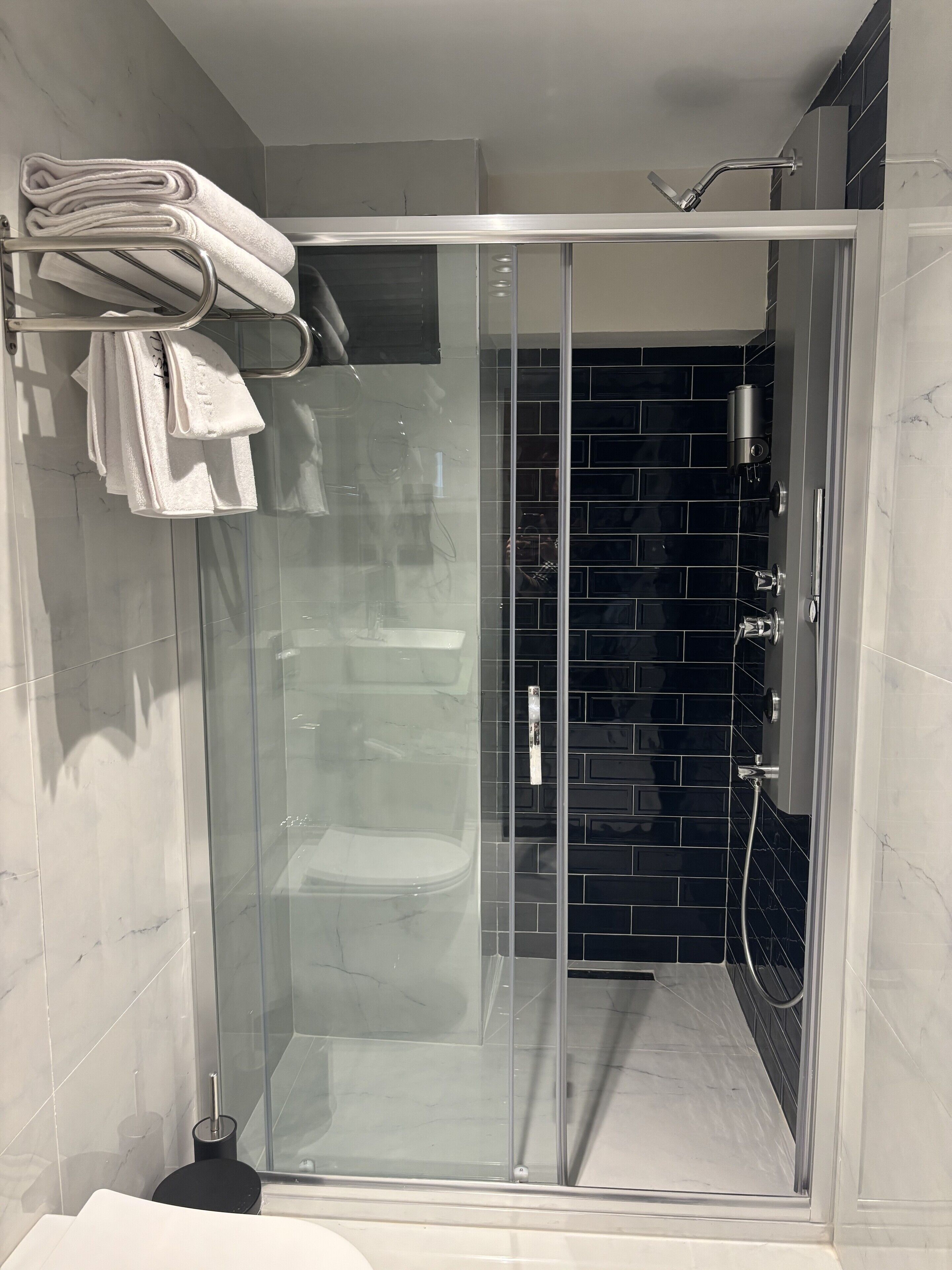 A modern bathroom with a glass-enclosed shower, black tiled walls, and neatly stacked towels on a metal rack.