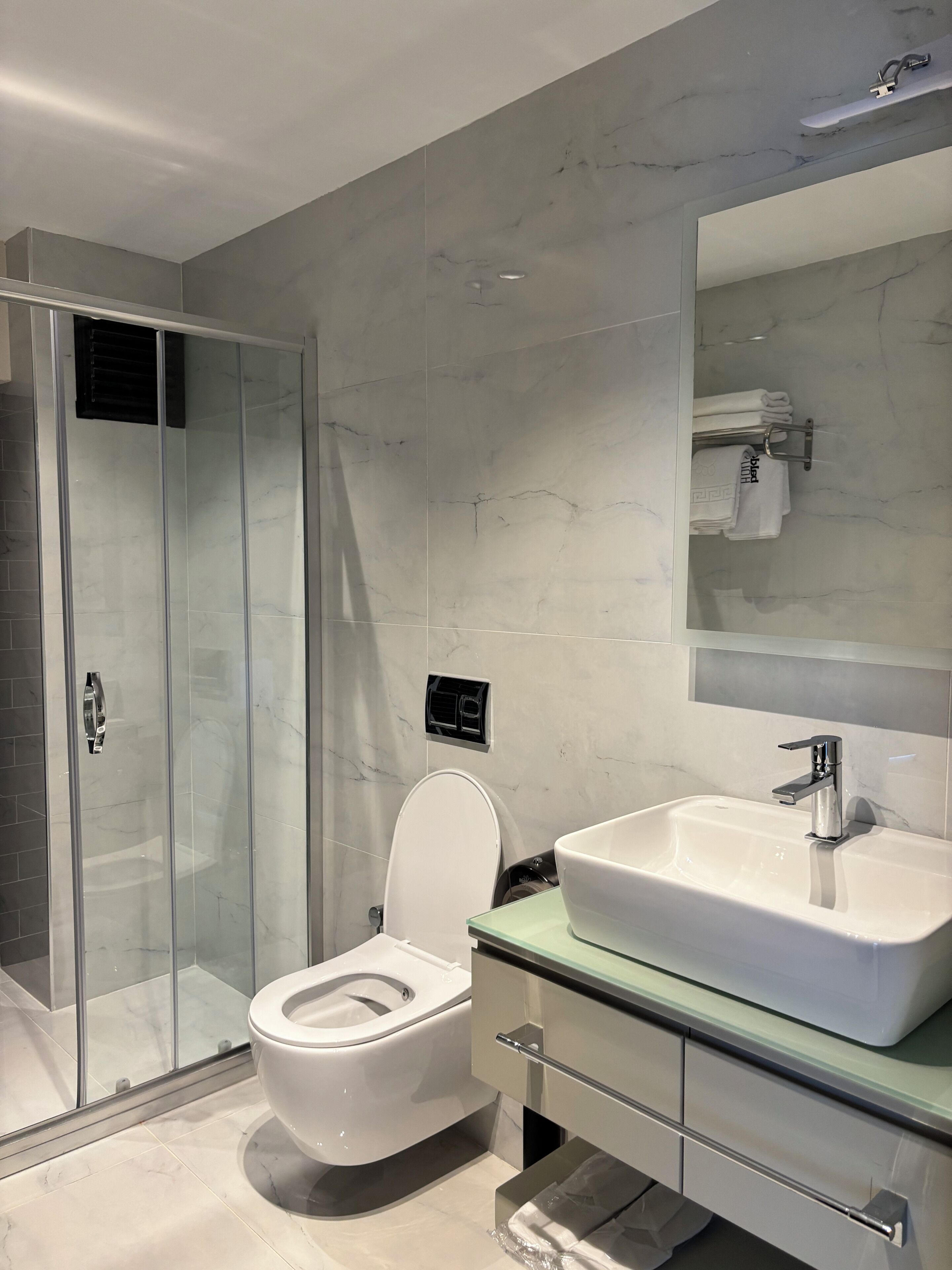 Un baño moderno con cabina de ducha de vidrio, inodoro blanco, espejo grande y lavamanos con encimera de vidrio.