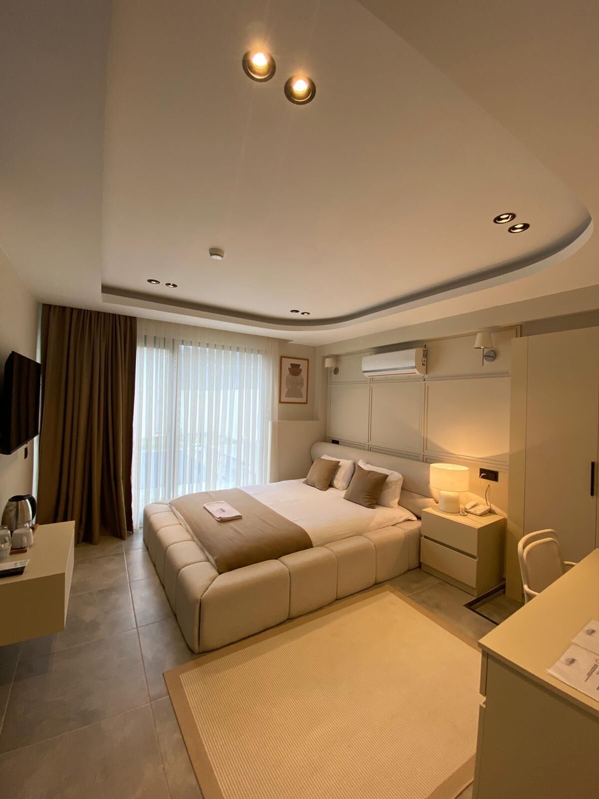 A modern hotel room with a large bed, a bedside table with a lamp, a desk with a chair, and a large window with curtains.