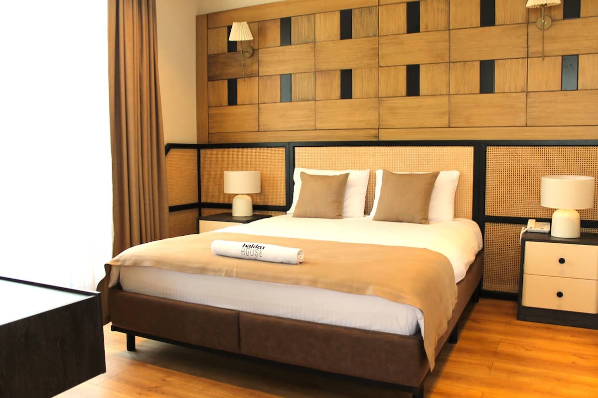 A hotel room with a large bed, two bedside tables, a headboard with a woven pattern, and a window with curtains.