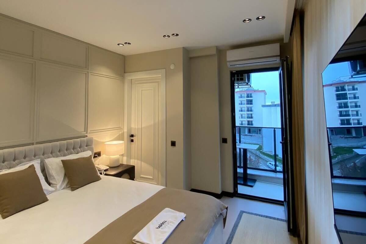 A hotel room with a large bed, a bedside table with a lamp, a door, and a view of the outside.