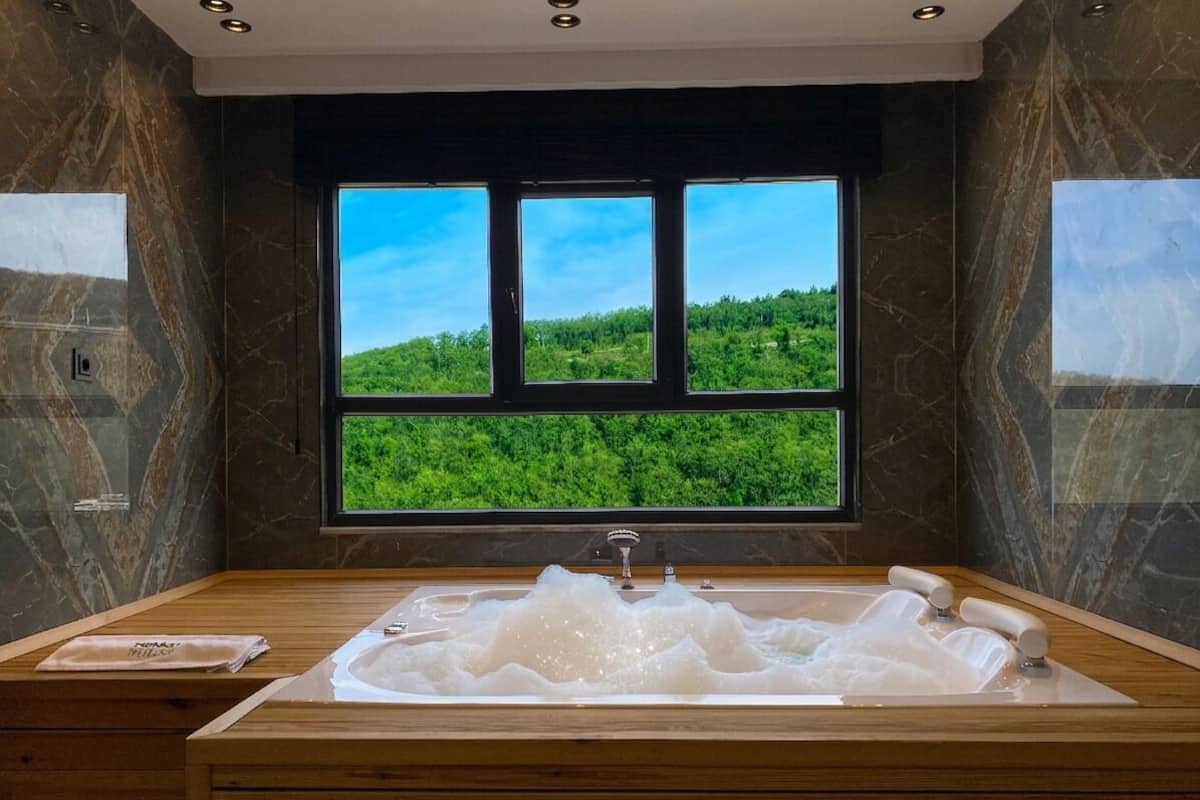 A modern bathroom with a large bathtub, a wooden vanity, and a window offering a view of a green landscape.