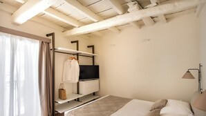 Quarto com cama, televisão, prateleira com uma mala e luminária.